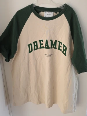 Dreamer t-shirt från Axel Arigato - Snygg t-shirt från Axel Arigato med beige framsida och mörkgröna ärmar. Trycket 'DREAMER' i grönt framtill ger en cool retrovibe. Klassisk raglanmodell med kontrastfärgade detaljer. Perfekt för en avslappnad stil. Passar s och m. Nypris 500-600kr