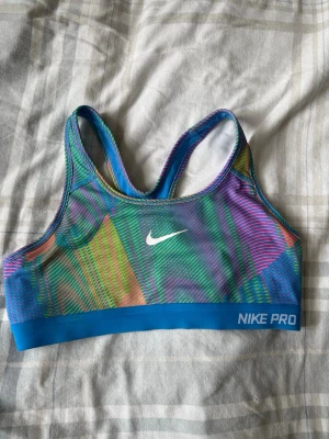 Nike Pro färgglad sport-bh - Nike Pro sport-bh i färgglatt mönster med blå, grön, lila och orangea toner. Tillverkad i Dri-FIT-material som andas och transporterar bort svett. Brottarrygg och elastisk nederkant med Nike Pro-logga. Perfekt för träning och sport.