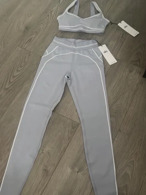 Grå set från Alo med vita detaljer - Snyggt ljusblått set från Alo med sport-bh och matchande tights. Setet har vita kontrastsömmar och en modern, sportig look. Materialet är stretchigt och följsamt, perfekt för en aktiv livsstil. Både bh och tights har diskreta Alo-loggor framtill. Är tillgänglig i alla storlekar och det kommer som ett sett !! 