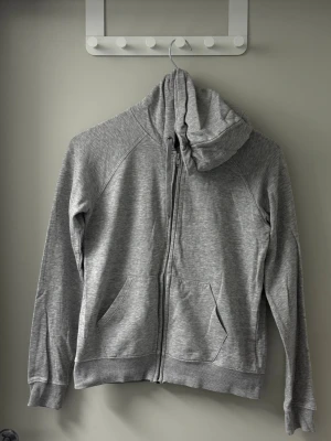 Grå zip-hoodie med fickor - En klassisk grå hoodie med dragkedja och två stora framfickor. Tröjan har huva och ribbade muddar vid ärmslut och nederkant. Perfekt för en avslappnad stil och enkel att matcha med andra plagg.