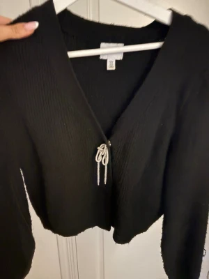 Svart kofta med dragkedja från Gina Tricot - Snygg svart kofta från Gina Tricot med dragkedja framtill och dekorativ knutdetalj. Koftan har v-ringning, lång ärm och är gjord i ett mjukt, stickat material. Perfekt att slänga över en topp för en chill look. M men sitter som S