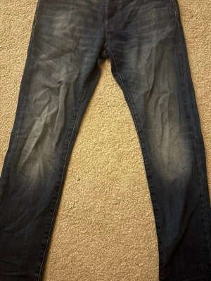 Mörkblå slim fit jeans från g-starraw - Säljer ett par mörkblå slim fit jeans från H&M med klassisk femficksdesign och snygga slitningar på låren. Jeansen har normal midja och smala ben, perfekta för en trendig och avslappnad look. Tillverkade i stretchigt denim för extra komfort.