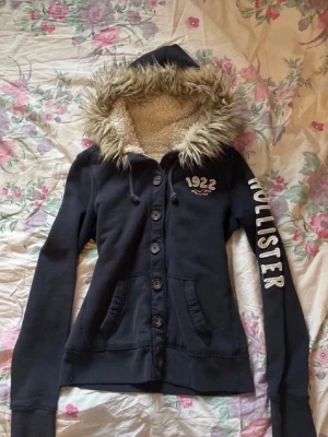 Hollister hoodie med knappar - En vintage Hollister hoodie med knappar och päls i storlek M, men skulle säga passar också som en S. Färgen är en mörk marinblå, snygga detaljer och text. Defekter förekommer då den är använd och tvättad flera gånger, exempel: trådar, urtvättad osv. Skicka gärna prisförslag🌷