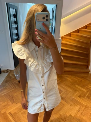 Vit kort klänning med volang - Säljer en vit kort klänning med stora volangdetaljer över axlarna och knappar framtill. Klänningen har skjortkrage och är ärmlös, perfekt för en trendig och somrig look. Materialet känns som bomull och passformen är normal.