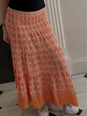 Mönstrad långkjol i orange och beige - Säljer en flowy långkjol med bohemiskt mönster i orange och beige toner. Kjolen har smock i midjan och ett lätt, luftigt tyg som passar perfekt till varma dagar. Den har volangdetaljer och ett färgglatt print som ger en chill vibe.