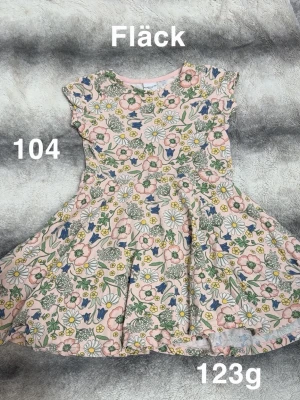 Blommig kortklänning med volang - Söt kortklänning med blommigt mönster i rosa, blått, grönt och gult. Klänningen har volangdetaljer vid axlarna och en rund halsringning. Perfekt för sommarens alla tillfällen.🌸 En fläck. Storlek 104