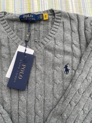 Grå kabelstickad tröja Polo Ralph Lauren - Snygg grå kabelstickad tröja från Polo Ralph Lauren med V-ringning och klassisk marinblå logga broderad på bröstet. Tröjan har långa ärmar och ribbade muddar vid ärmslut och nederkant. Perfekt för en stilren och avslappnad look.