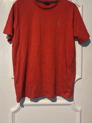 Röd t-shirt från Gant - En klassisk röd t-shirt från Gant med rund halsringning och diskret logga på bröstet. T-shirten har korta ärmar och är tillverkad i mjuk bomull, perfekt för en avslappnad och stilren look.