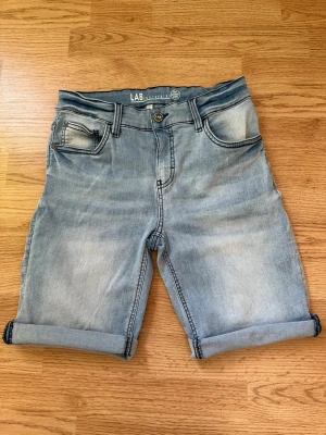 Ljusblå denimshorts från LAB Industries - Säljer ett par ljusblå denimshorts från LAB Industries med klassisk femficksdesign. Shortsen har normal passform och stängs med knapp och dragkedja. Perfekta för varma dagar och enkel att matcha med olika stilar.