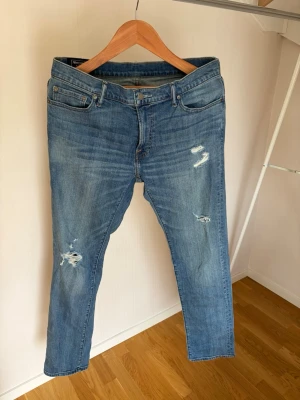 Blå slitna jeans med raka ben - Säljer ett par ljusblå jeans med raka ben och slitningar på framsidan. Jeansen har klassisk femficksmodell, normal midja och är tillverkade i denim. Perfekta för en avslappnad och trendig look.