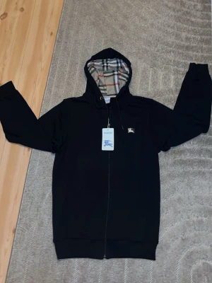 Svart Burberry hoodie med dragkedja - Svart hoodie från Burberry med dragkedja och klassiskt rutigt foder i huvan. Ikoniska Burberry-loggan broderad på bröstet. Mjuk bomullskänsla och justerbar huva med snören. Perfekt för en stilren och avslappnad look.