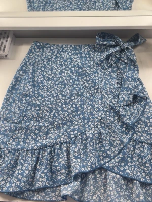 Blå blommig omlottkjol med volang - Supersöt blå omlottkjol med vitt blommönster och volangkant. Kjolen har en knytning i midjan som ger en snygg detalj och passar perfekt till varmare dagar. Lätt och luftig känsla. Dragkedja finns även så att det är lätt att sätta på sig. Djur finns i hemmet, tvättas innan leverans.💗