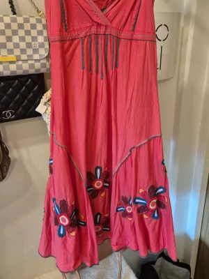 Röd klänning med broderi från Desigual - Färgstark röd klänning från Desigual med broderade blommor i flera färger nedtill. Klänningen har omlott-ringning, dekorativa sömmar och snörningsdetaljer framtill. Små guldiga kulor fastsydda i blommorna. Tillverkad i ett lätt bomullstyg, perfekt för varma dagar.