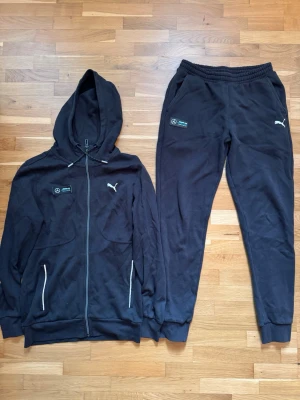 Svart Tracksuit från Puma x Mercedes - Svart tracksuit från Puma i samarbete med Mercedes AMG Petronas. Dragkedja framtill, två fickor med vita detaljer och broderade loggor på bröstet. Tillverkad i mjukt bomullsmaterial och har en klassisk huva med snören. Perfekt för dig som gillar sportig och clean stil.