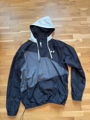 Under Armour svart/vit vindjacka - Snygg vindjacka från Under Armour i svart och vitt med stor huva och dragsko. Jackan har en cool grafisk logga på ryggen och Under Armour-logga på bröstet. Tillverkad i lätt, vindtätt material och har elastiska muddar vid ärmar och midja.