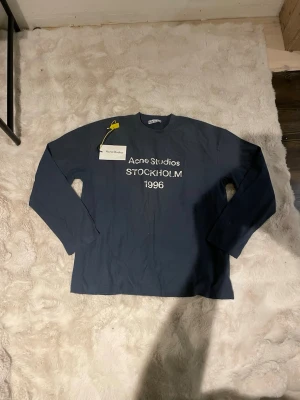 Blå långärmad Acne Studios t-shirt - Snygg blå långärmad t-shirt från Acne Studios med tryck 'STOCKHOLM 1996' på bröstet. Klassisk rund halsringning och relaxed fit. Perfekt för dig som gillar stilrena plagg med en urban touch. Den passar även M