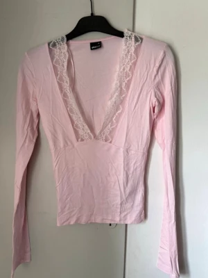 Ljusrosa topp med spets från Gina Tricot - Supersöt ljusrosa långärmad topp från Gina Tricot med djup v-ringning och spetsdetaljer längs urringningen. Toppen är figurnära och har en mjuk, stretchig känsla som passar perfekt till jeans eller kjol.
