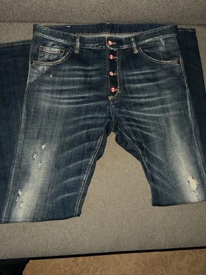 DSQUARED2 slitna blå jeans - Säljer ett par blå jeans från DSQUARED2 med slitningar och snyggt tvättad look. Jeansen har röda knappar med märkets bokstäver och klassisk femficksmodell. Materialet är denim i bomull och passformen är rak/slim.