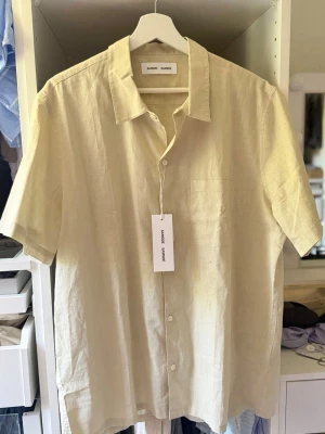 Beige linneskjorta från Samsoe Samsoe - Säljer en beige kortärmad skjorta från Samsoe Samsoe i luftigt linne. Skjortan har klassisk krage, bröstficka och knappar framtill. Perfekt för varma dagar och enkel att styla till olika outfits.