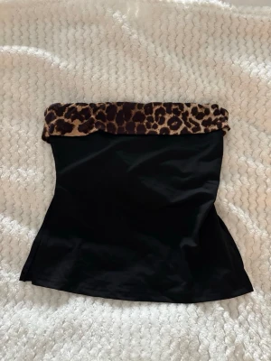 Svart tubtopp med leopardkant från SHEIN - Säljer en svart tubtopp från SHEIN med en snygg leopardmönstrad kant upptill. Toppen är axelbandslös och har en tight passform som framhäver figuren. Perfekt för dig som vill sticka ut lite extra.