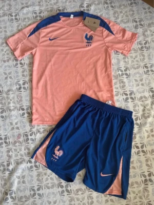 Nike fotbollsträningsset, två färger - Två olika fotbollsträningsset från Nike. Ena setet är ljusrosa med blå detaljer och FFF-logga, det andra är svart med rosa detaljer och Chelsea FC-logga samt trivago-tryck. Båda seten består av t-shirt och shorts i lätt, snabbtorkande material.