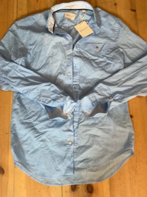 Ljusblå skjorta med knappar - Snygg ljusblå skjorta med klassisk krage och vita knappar. Skjortan har lång ärm och diskreta ränder på insidan av manschetterna. Perfekt till jeans eller chinos för en clean look.