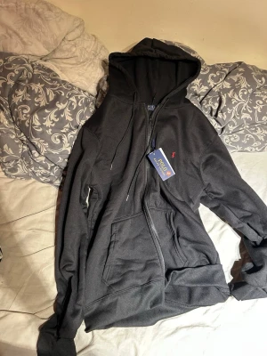 Svart hoodie från Polo Ralph Lauren - Snygg svart hoodie från Polo Ralph Lauren med dragkedja framtill och klassisk röd logga broderad på bröstet. Hoodien har huva med dragsko och stora fickor framtill. Tillverkad i mjukt bomullsmaterial som känns skönt mot huden. Perfekt för en avslappnad och stilren look.