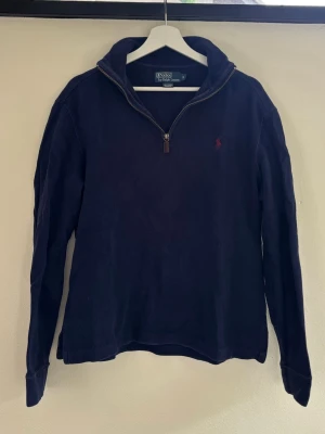 Mörkblå halvzip från Polo Ralph Lauren - Mörkblå långärmad tröja från Polo Ralph Lauren med halvzip och hög krage. Tröjan har det klassiska röda Polo-logot broderat på bröstet och är tillverkad i ett mjukt bomullsmaterial. Perfekt för en avslappnad men stilren look.
