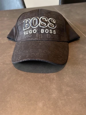 Svart keps från Hugo Boss - Snygg svart keps från Hugo Boss med stor vit broderad BOSS-logga framtill och diskret mönster med mindre logotyper över hela kepsen. Justerbar rem baktill med metallspänne. Klassisk böjd skärm och stilren design, perfekt för en trendig look.