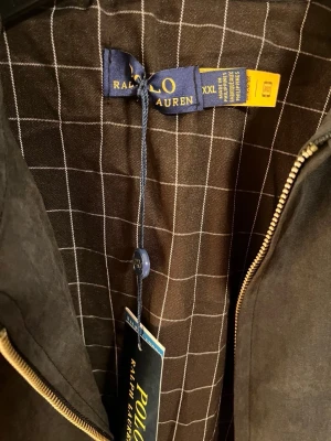 Svart jacka från Polo Ralph Lauren - Snygg svart jacka från Polo Ralph Lauren i storlek XXL. Jackan har klassisk krage, dragkedja framtill och ett rutigt innerfoder i brunt och vitt. Materialet ser ut att vara mockaliknande och jackan har en broderad logga på bröstet.