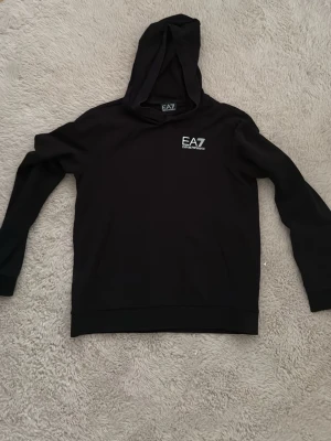 Svart hoodie från EA7 Emporio Armani - Snygg svart hoodie från EA7 Emporio Armani med liten vit logga på bröstet. Klassisk modell med huva och långa ärmar. Tillverkad i mjukt material som känns skönt mot huden. Perfekt för en avslappnad och trendig look.
