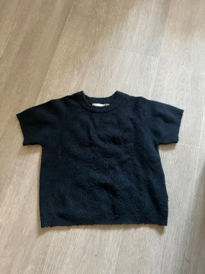 Svart stickad t-shirt - En svart stickad t-shirt med korta ärmar och rund hals. Tröjan har en enkel och stilren design utan synliga mönster eller detaljer. Perfekt för dig som gillar minimalistisk stil och vill ha något bekvämt och mjukt i garderoben. Använd några få gånger!