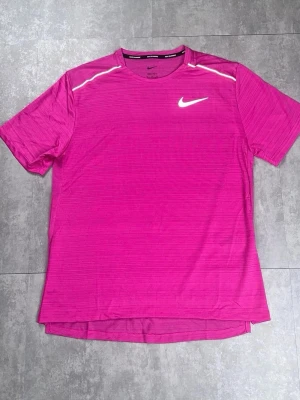 Rosa sporttröja från Nike - Snygg rosa sporttröja från Nike med rund halsringning och korta ärmar. Tröjan har diskreta horisontella ränder och en vit Nike-logga på bröstet samt vita detaljer vid axlarna. Perfekt för träning eller löpning.