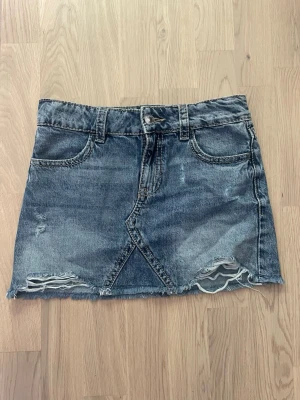 Blå jeanskjol med slitningar - Snygg blå jeanskjol i kort modell med slitna detaljer och rå kant nertill. Kjolen har fem fickor, bälteshällor och knappstängning fram. Perfekt för en avslappnad och trendig look.