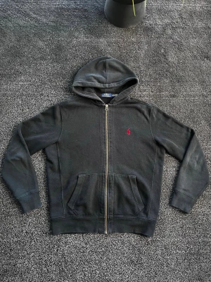 Ralph Lauren zip Hoodie  - Svart hoodie med dragkedja från Polo Ralph Lauren. Klassisk design med huva, kängurufickor och röd broderad logga på bröstet. Tillverkad i mjukt bomullsmaterial som är skönt att ha på sig. Perfekt för en avslappnad och stilren look. Skick 8/10 saknar snören, storlek L men passar mer som M. Pris går att diskutera✅