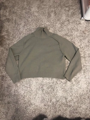 Grön stickad polotröja - Mysig grön stickad polotröja med lång ärm och ribbade muddar vid ärmslut och nederkant. Tröjan har en hög, bred polokrage och är rak i modellen. Perfekt för kyliga dagar och enkel att matcha med olika outfits.