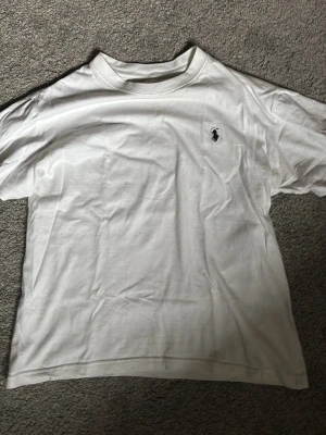 Vit t-shirt från Ralph Lauren - Klassisk vit t-shirt från Ralph Lauren med den ikoniska broderade loggan på bröstet. T-shirten har rund hals och korta ärmar, tillverkad i mjuk bomull för en skön känsla. Perfekt basplagg som funkar till allt.