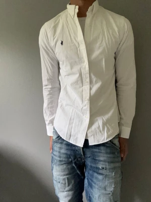 Vit skjorta från Ralph Lauren - Klassisk vit skjorta från Ralph Lauren med button-down krage och broderad marinblå logga på bröstet. Skjortan har lång ärm, knappar framtill och är tillverkad i bomull. Perfekt till jeans eller chinos för en clean och stilren look.