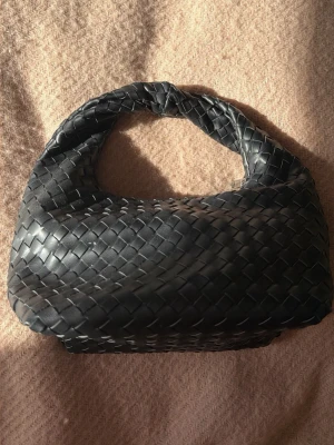 Svart flätad handväska Gina Tricot - Snygg slutsåld svart handväska från Gina Tricot med flätad design och mjuk struktur. Väskan har ett kort handtag och stängs med dragkedja. Perfekt accessoar för att lyfta din outfit med en trendig touch. Som ny.