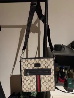 G väska - Snygg beige axelväska  med klassiskt GG-monogram, svart justerbar axelrem och detaljer i svart läder. Väskan har en framficka med dragkedja och den ikoniska gröna och röda randen framtill. Perfekt för dig som vill ha en trendig och lyxig accessoar.