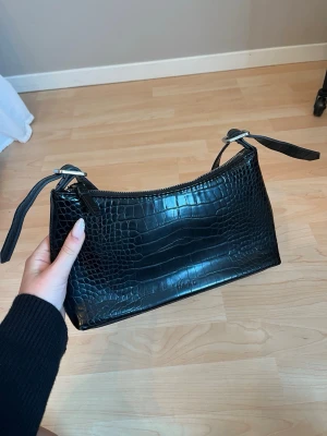 Väska - Snygg svart handväska i croco-mönstrat konstläder med silverfärgade detaljer och dragkedja upptill. Har några skavanker som syns på bilderna. 