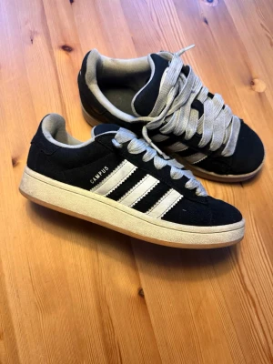 Svarta Adidas Campus sneakers - Säljer ett par klassiska Adidas Campus sneakers i svart mocka med vita detaljer och de ikoniska tre ränderna på sidan. Skorna har gråa skosnören och en vit sula med gummiyta. Perfekta för dig som gillar retrostil och streetwear. Detta par är i storlek 39 men passar upp mot 40. Snörerna är lite sämre skick en skorna pågrund av smutsen men det är lätt att fixa genom att tvätta dem rätt! Hör gärna av er vid frågor eller prisförslag😃✅.