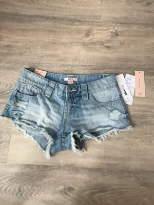  jeansshorts - Säljer ett par ljusblå jeansshorts från Ginatricot  med råa kanter och slitna detaljer. Shortsen har fyra fickor, bälteshällor och en klassisk knappgylf. Perfekta för varma dagar och ger en avslappnad vibe.