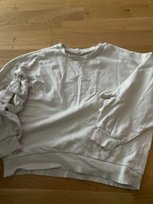 Vit sweatshirt med knyt på ärmen - Säljer en stilren vit sweatshirt med rund hals och ribbade muddar. Tröjan har en unik detalj med knyt på ena ärmen som ger en cool twist. Tillverkad i mjukt bomullsmaterial och har en avslappnad passform. Perfekt för dig som gillar enkla men snygga plagg.
