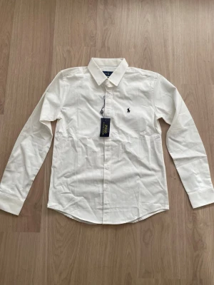 Vit skjorta från Polo Ralph Lauren - Klassisk vit skjorta från Polo Ralph Lauren med lång ärm och knappstängning framtill. Skjortan har en broderad logga på bröstet och är tillverkad i mjuk bomull. Perfekt till både jeans och chinos för en clean look.