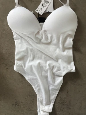 Vit vadderad body från Gina Tricot - Säljer en vit body från Gina Tricot med push-up kupor och justerbara axelband. Bodyn har en djup urringning och stringmodell baktill. Tillverkad i mjukt stretchigt material för en bekväm passform. Perfekt att styla under skjortor eller tröjor. Helt oanvänd och prislap kvar på. 