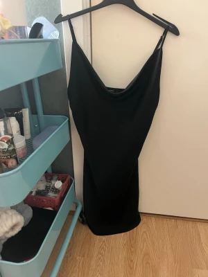 Svart slipklänning med tunna band - Säljer en svart, enkel och stilren slipklänning med tunna axelband och lätt glansigt tyg. Klänningen har en smickrande v-ringning och är kort i modellen, perfekt för en trendig look. Materialet känns mjukt och följsamt mot huden.