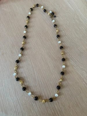 Halsband med svarta och vita pärlor - Snyggt halsband med svarta och vita pärlor samt guldfärgade detaljer. Pärlorna är jämnt fördelade längs en tunn guldfärgad kedja som ger ett elegant och trendigt intryck. Perfekt accessoar för att lyfta din outfit med en stilren touch.
