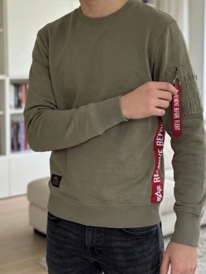 Olivgrön Alpha Industries sweatshirt - Cool olivgrön sweatshirt från Alpha Industries med röda detaljer och texten 'Remove Before Flight' längs sidorna och på ärmfickan. Tröjan har rund hals, ribbade muddar och en ficka med dragkedja på ena ärmen. Perfekt för en streetwear-look.