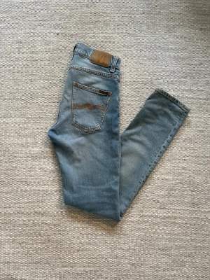 Ljusblå jeans från Nudie Jeans - Säljer ett par ljusblå jeans från Nudie Jeans med klassisk femficksdesign och bruna sömmar. Jeansen har raka ben och är tillverkade i mjuk denim. Snygg detalj med Nudie-loggan på bakfickan och läderpatch i midjan.